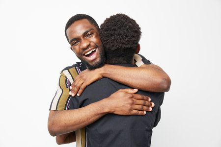Two black men sharing a joyful embrace, surrounded by a bright white background. Their vibrant clothing enhances the positive atmosphere of the moment.の写真素材