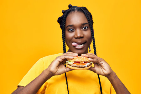 Young Black woman smiling and holding a delicious burger against a vibrant yellow background,...