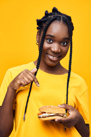 Smiling young African girl pointing at a delicious burger, wearing a bright yellow shirt against a vibrant yellow background, conveying a positive and cheerful vibeの写真素材