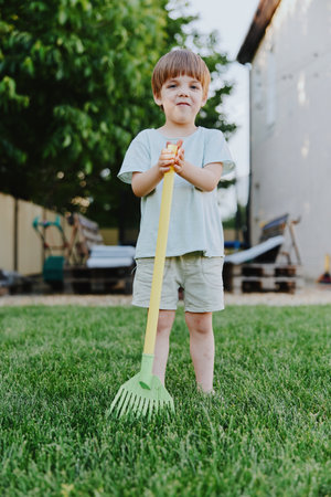 Young boy holding a colorful rake, standing on lush green grass, smiling with joy. This playful moment captures the essence of childhood and outdoor fun.の写真素材