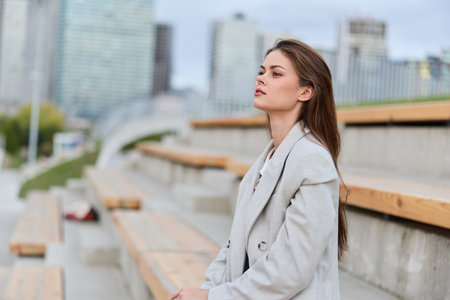Woman in light coat sitting outdoors on urban wooden bleachers contemplating with cityscape background, autumn casual fashion and pensive moodの写真素材