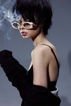 stylish woman with short dark hair wearing sunglasses and black clothing blowing smoke in moody lighting with a neutral backgroundの写真素材