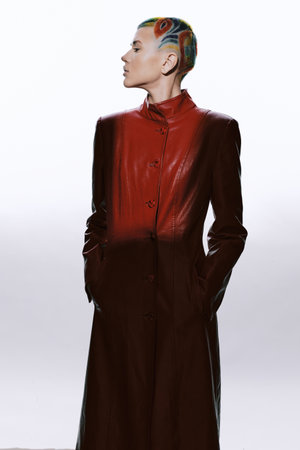 Woman with colorful shaved hair wearing a long red leather coat stands confidently against a white...