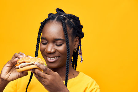 Cheerful young woman enjoying a delicious burger against a vibrant yellow background, showcasing a...
