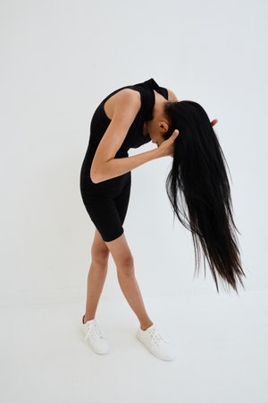 slender woman with long black hair in black sleeveless outfit bending forward against plain white...