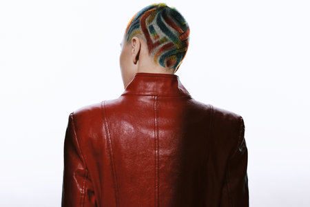 Woman with colorful shaved hairstyle wearing a glossy red leather jacket, viewed from behind against a bright white background, showcasing edgy modern fashion style.の写真素材