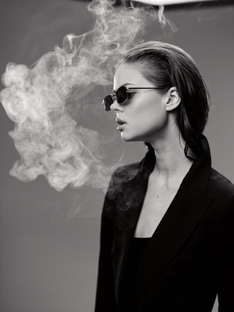 black and white woman wearing sunglasses exhaling dense smoke with stylish slicked-back hair and dark clothing in dramatic lighting and neutral backgroundの写真素材