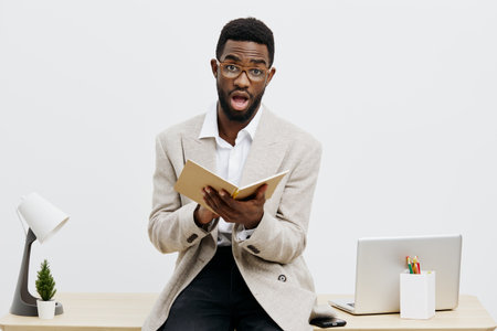 Professional portrait of a surprised man with glasses and dark skin, reading a book at his desk, wearing a beige blazer, minimal office environment, bright natural lighting, focused expression, indoor shot. People conceptの写真素材