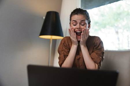 Excited woman in a brown shirt reacting joyfully while using a laptop at home, expressing happiness...