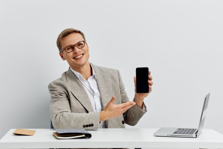Smiling man in office attire holding smartphone and showing it to the camera, sitting at a desk with...