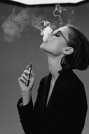 black and white woman vaping with glasses exhaling smoke against plain background in stylish outfit,...