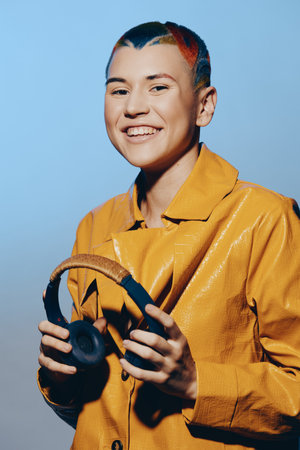 Smiling woman with multicolored hair wearing a bright yellow jacket holds black headphones against a...