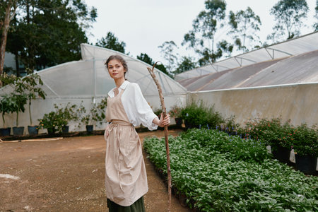 Young woman in a garden wearing a white shirt and apron, holding a stick among lush greenery, showcasing sustainable gardening and a serene atmosphereの写真素材