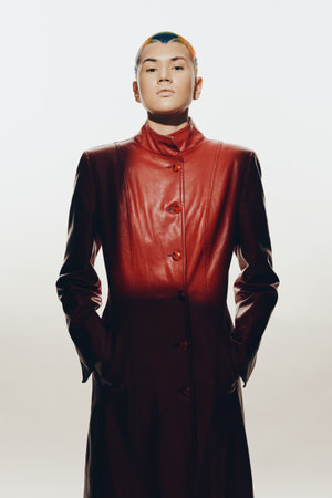 woman in long red leather coat with short hair stands confidently against white background,...