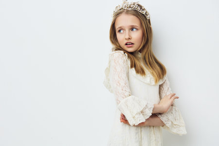 A young girl wearing a white lace dress and tiara, expressing a thoughtful emotion against a soft white background, ideal for concepts of innocence and childhood dreamsの写真素材