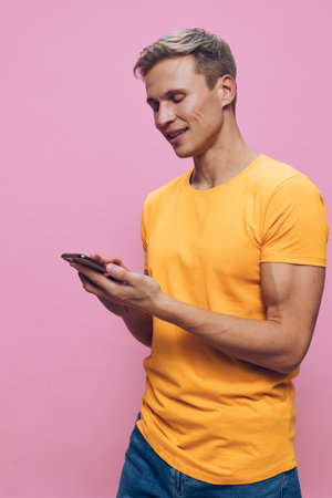 Man in casual yellow t-shirt holding and looking at mobile phone isolated on pink background studio...