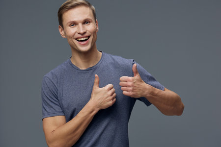 smiling happy young caucasian man wear blue t-shirt look camera show thumb up gesture isolated on grey background digital detox concept mental health and gadgetsの写真素材