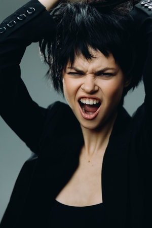 angry woman with short black hair expresses frustration wearing black jacket with open mouth and...