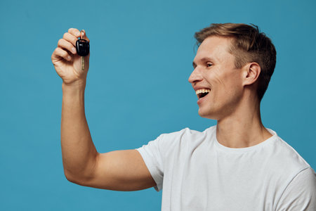 Smiling young man with short hair wearing a plain white t-shirt holding a car key up towards the camera isolated on a solid blue background studio portrait. People lifestyle concept with excitement and happinessの写真素材