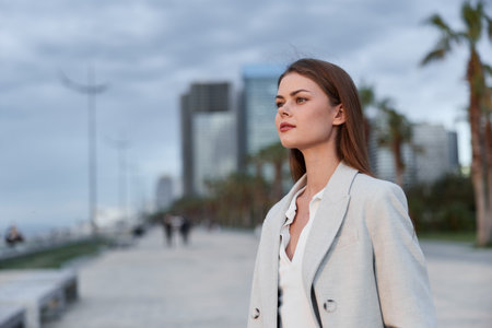Business woman in light coat walking outdoors in city with serious confident expression and modern buildings backgroundの写真素材