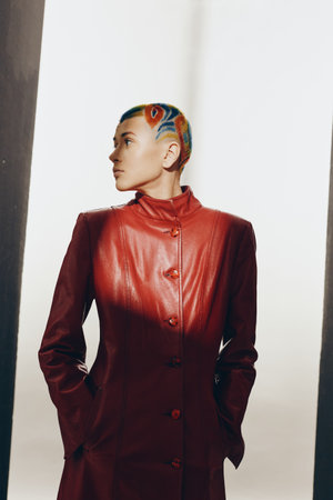 Young woman with colorful short hair wearing red leather coat stands confidently against bright...