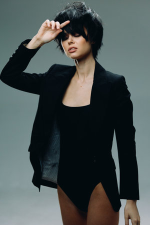 Stylish woman in black bodysuit and blazer posing with hand near face against gray background.