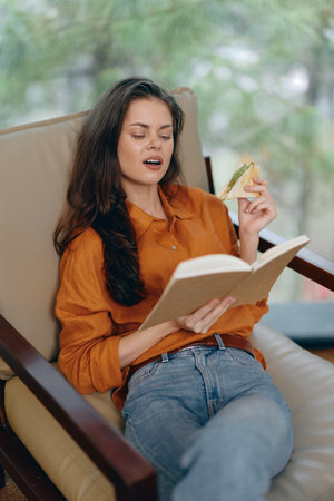 Young woman relaxing with a book and sandwich, wearing a cozy orange shirt, enjoying a moment of...