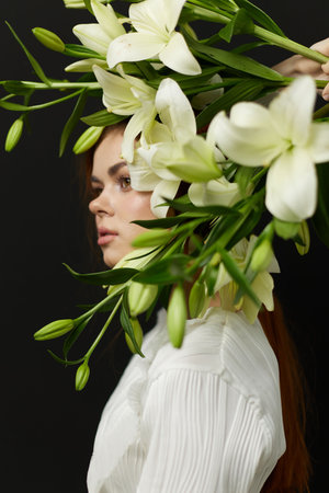 Elegant young woman with long brown hair adorned with white lilies, showcasing a striking floral crown, exuding serenity and beauty against a dark backgroundの写真素材