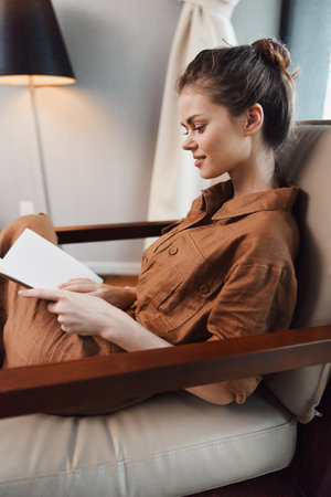 A young woman with brown hair sitting comfortably in a chair, reading a book in a cozy setting with...
