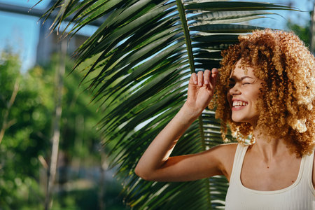 smiling woman with curly hair enjoys sunlight under palm leaves during tropical summer day, natural...