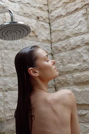young woman under outdoor shower with wet hair, relaxed posture, natural stone wall background,...