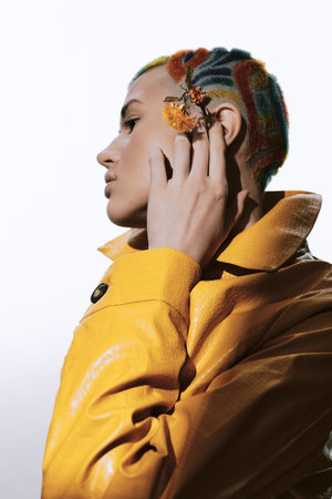Woman with colorful buzzcut hairstyle and yellow jacket posing with dried flowers near ear, showcasing unique fashion style against plain white background.の写真素材