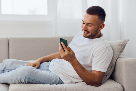 Young man relaxing on a couch, smiling while using a smartphone in a bright, cozy living room,...