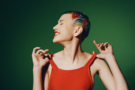 Young woman with colorful shaved hair and red dress smiling joyfully against a green background,...