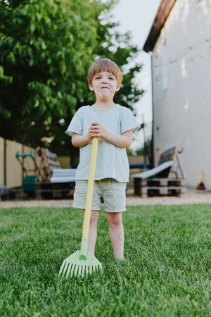 Happy boy holding a rake in a sunny garden, wearing casual clothes, enjoying outdoor activities and...