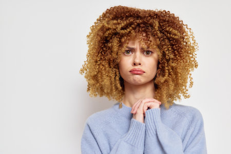 Sad woman with curly hair expressing disappointment, wearing a cozy blue sweater, set against a neutral background Emotional portrait capturing vulnerability and sensitivityの写真素材
