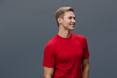 Smiling young man with blond hair wearing red t-shirt looking to the side against plain gray...