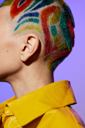 Woman with colorful painted hair and shaved sides wearing a bright yellow textured jacket against a vibrant purple background, showcasing creative self-expression and bold style.の写真素材