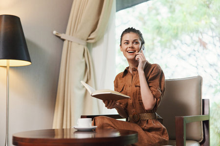 Smiling young woman speaking on the phone while reading a book in a cozy, well lit living space, embodying a relaxed lifestyle and enjoying leisure timeの写真素材