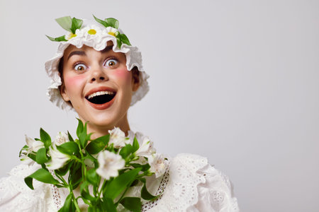 Cheerful young woman wearing a flower crown and white dress, holding a bouquet of flowers against a...