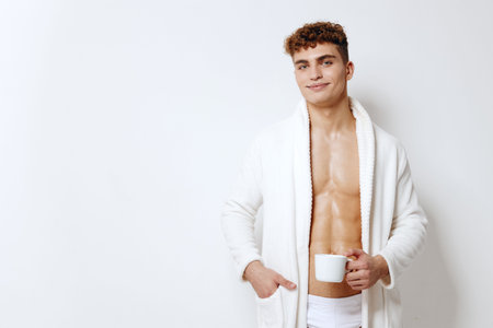 Confident young man in a white robe holding a mug, standing against a plain white background. Morning routine and self-care concept.の写真素材