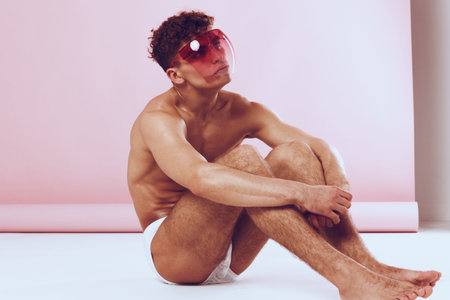 Stylish young man sitting on the floor, wearing bright sunglasses, showcasing a confident pose against a soft pink background. Fashion and self-expression concept.の写真素材