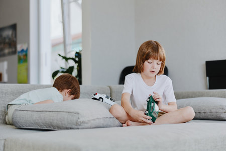 Two children playing indoors in a cozy living room, one boy focused on his toy while another boy explores the couch. Enjoying their afternoon together.の写真素材