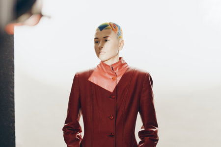 woman with colorful short hair wearing a red leather jacket standing in soft sunlight against a...