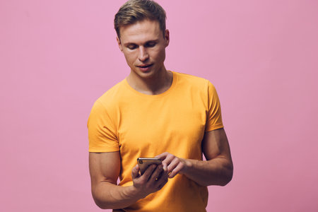 Smiling man with short hair wearing yellow t-shirt using mobile phone isolated on pink background. Casual lifestyle concept, young adult, communication, technology.の写真素材