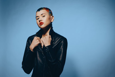 Young woman with short colorful hair and red lipstick wearing a black leather jacket posing confidently against a solid blue background in studio lighting.の写真素材