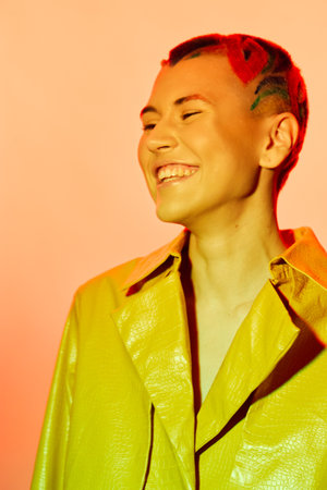 Smiling woman with bright red hair and a vibrant yellow jacket, radiating happiness and confidence against a warm colorful background, showing modern style and joy.の写真素材