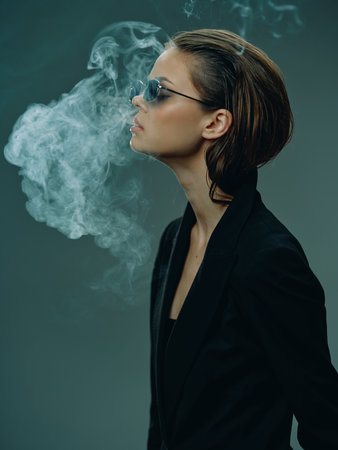 Profile of a stylish woman wearing sunglasses, exhaling smoke with a dark green backgroundの写真素材