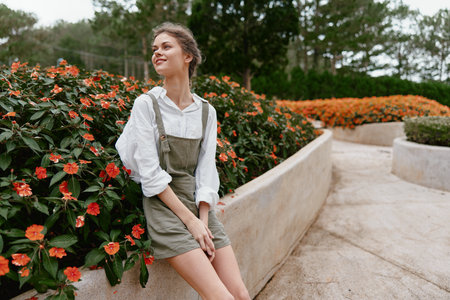 Young woman in green overalls with white shirt smiling near vibrant orange flowers in a sunny garden...