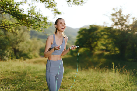 Young woman laughing while skipping rope in nature, showcasing fitness and joy in a sunny outdoor setting, perfect for healthy lifestyle themesの写真素材